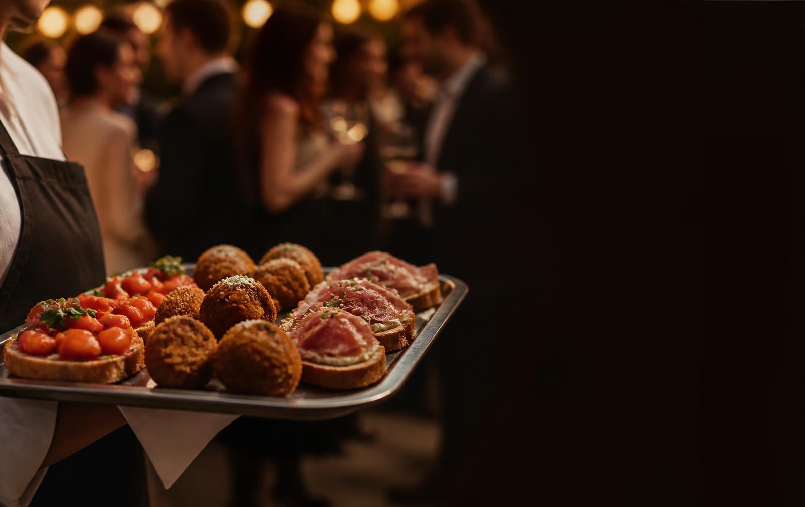 Server carrying a tray of passed bites — bruschetta, arancini and prosciutto crostini — through a reception